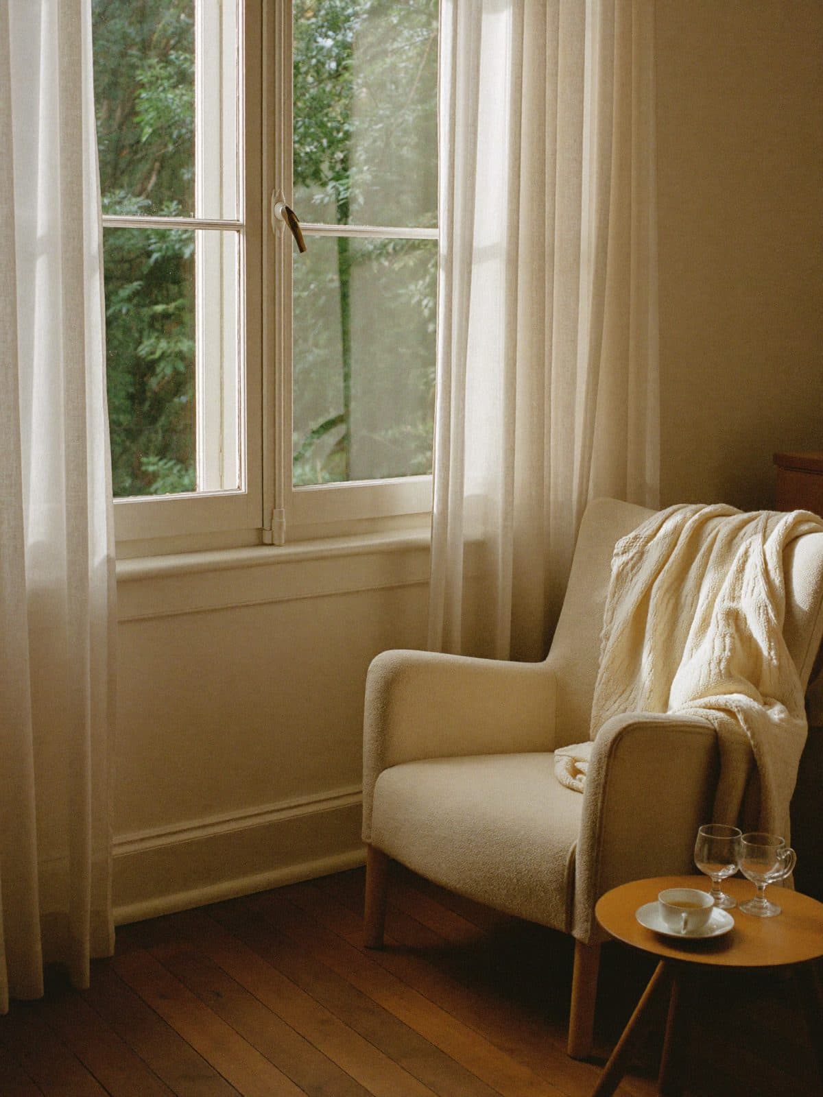 Ein heller Sessel mit Wolldecke am Fenster, daneben ein Tischchen mit einer Tasse Tee.